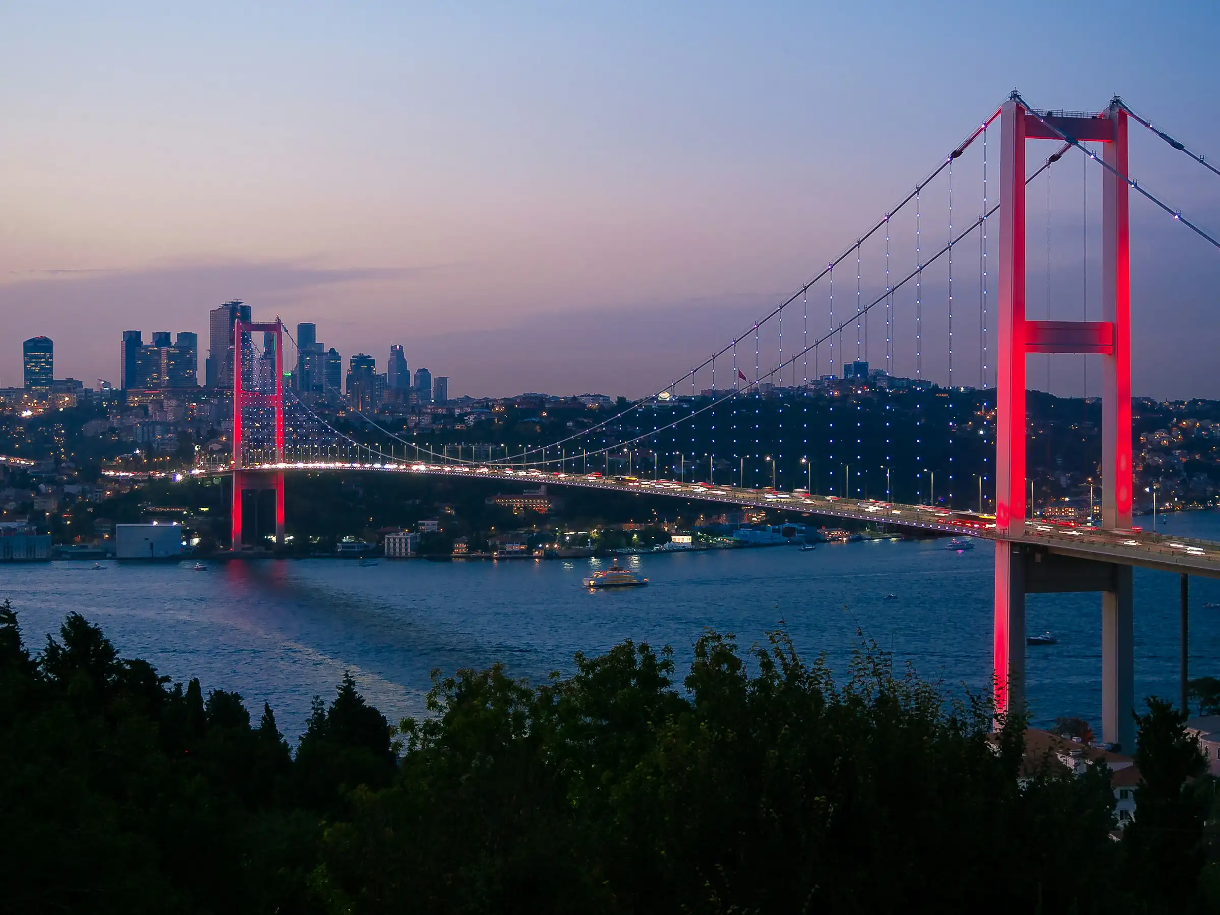 The Bosphorus Bridge in Istanbul at dusk, lit in red, with the city skyline behind